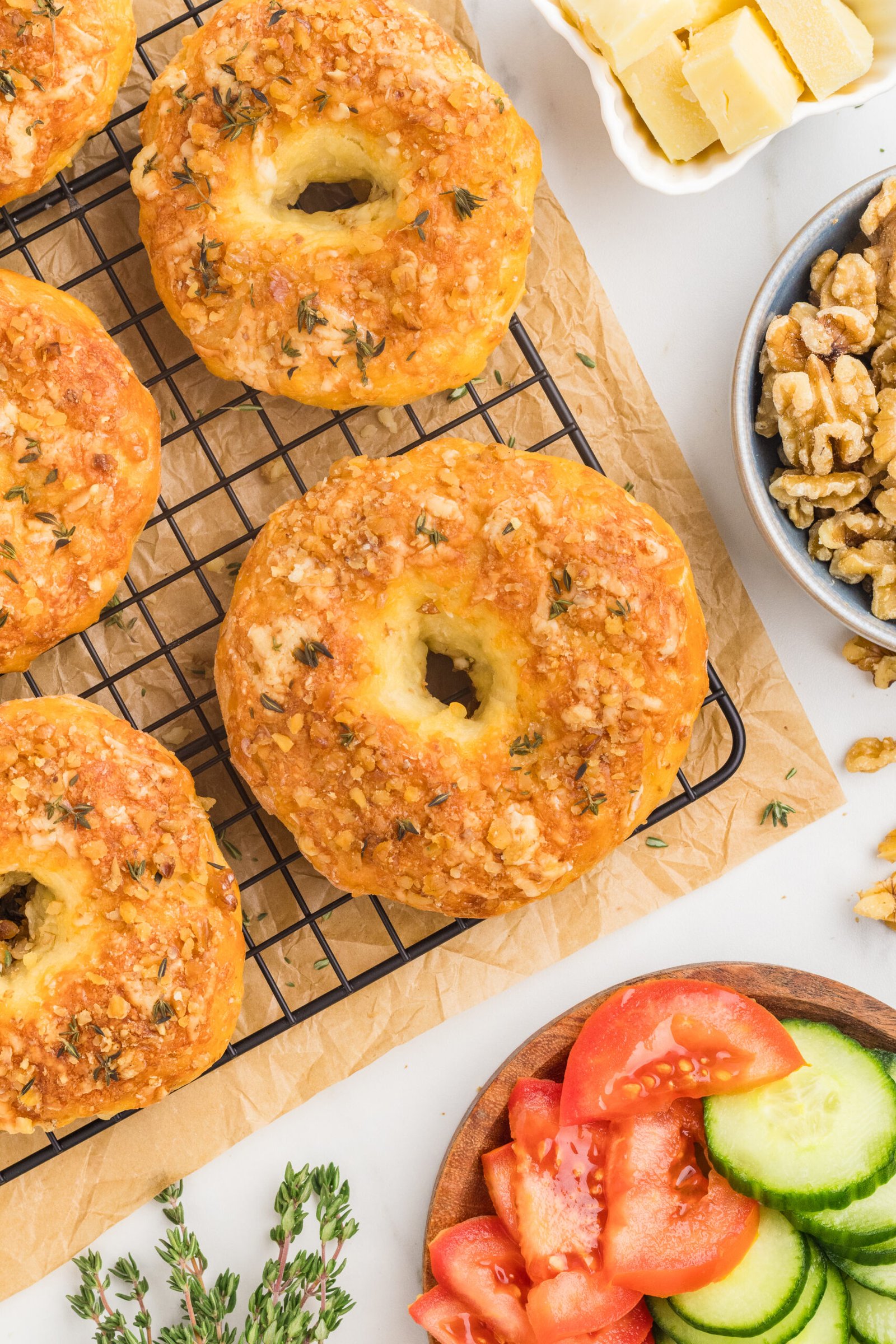 Overhead flat lay food photography of golden protein bagels on a wire cooling rack, styled with fresh breakfast sides like tomatoes and cucumbers to demonstrate balanced composition.