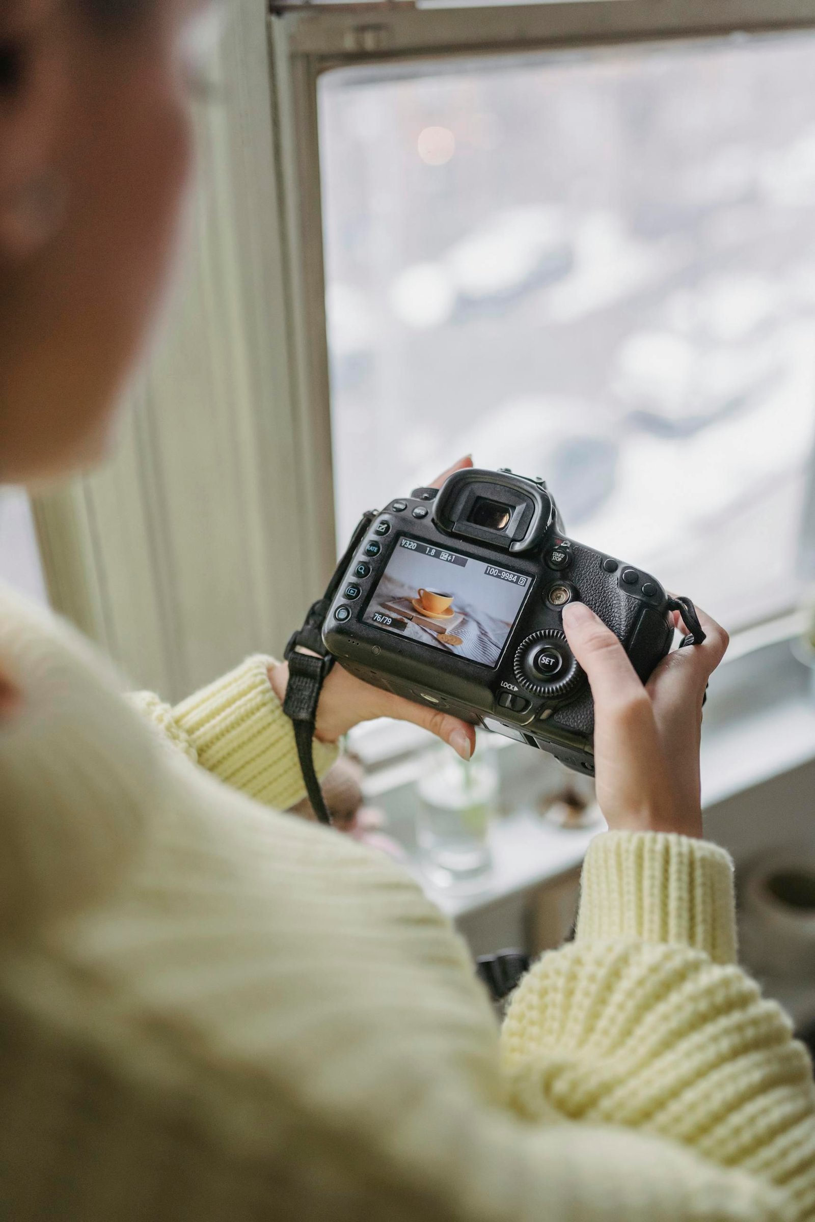 Female photographer checking a photo on a DSLR camera by a window, indoors.